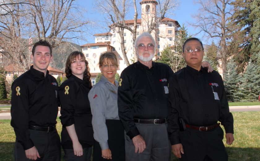 five people wearing grey and black standing, green trees in the background, beige and red building in the middle