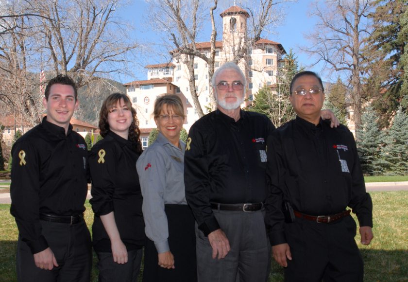 five people wearing grey and black standing, green trees in the background, beige and red building in the middle