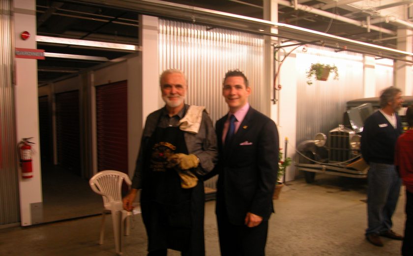 two men standing inside white and grey storage facility, one man wearing black apron, other man wearing black suit and pink dress shirt