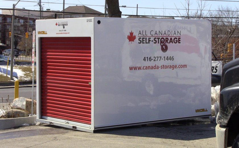 white storage unit with red door, sitting on grey pavement in parking lot, brown houses with grey roofs in the background