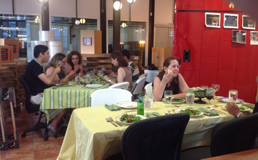 white and red coloured room with two long tables with a yellow and green table cloth, people sitting on black and white chairs eating food