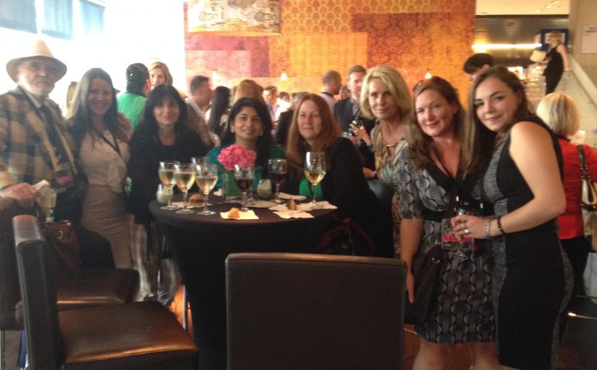 group photo eight women and one man, gathered around a black high table with black hair chairs, wine glasses on the table, red/orange/yellow wall