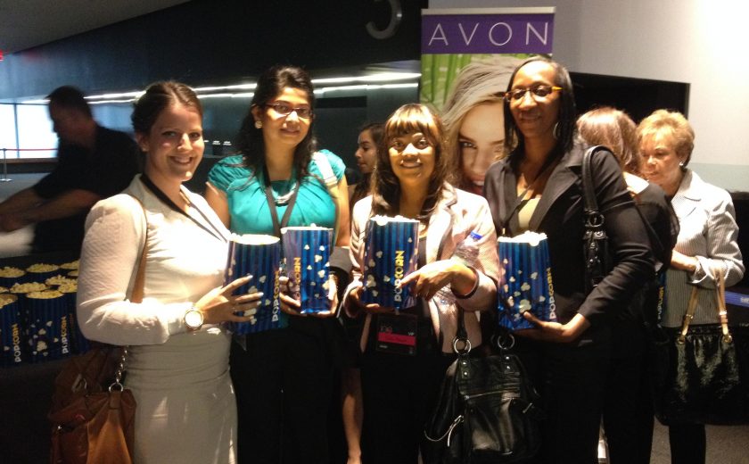 four women standing and smiling, holding blue and yellow bags of popcorn, inside a movie theatre