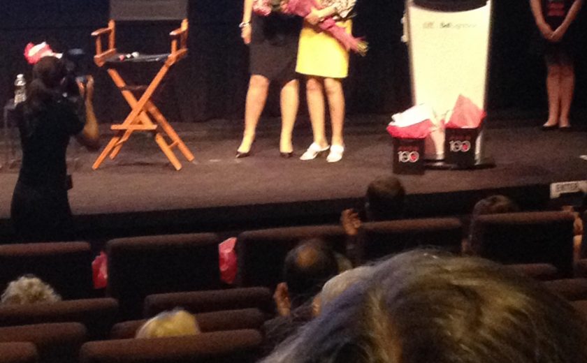 two women on stage holding a bouquet of pink flowers, woman in black taking a picture of them on stage