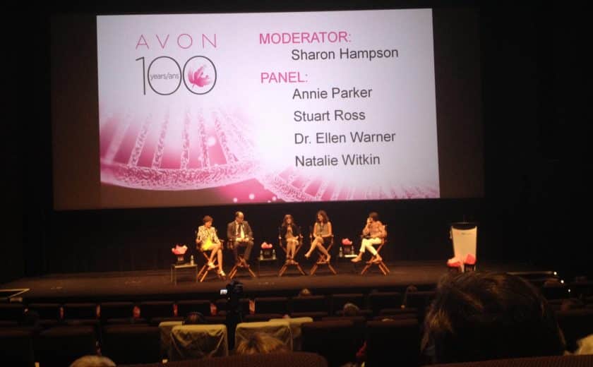 panel of five speakers sitting on brown chairs on stage, large screen with pink display