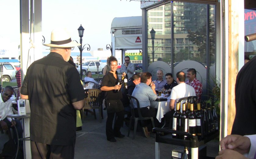 group of people sitting on the patio, cart full of black and gold alcohol bottles, parking lot filled with cars in the back, blue sky