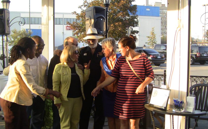 group of people gathered in front of sound speaker on the patio, blue sky, blue and white building in the background, parked cars in parking lot