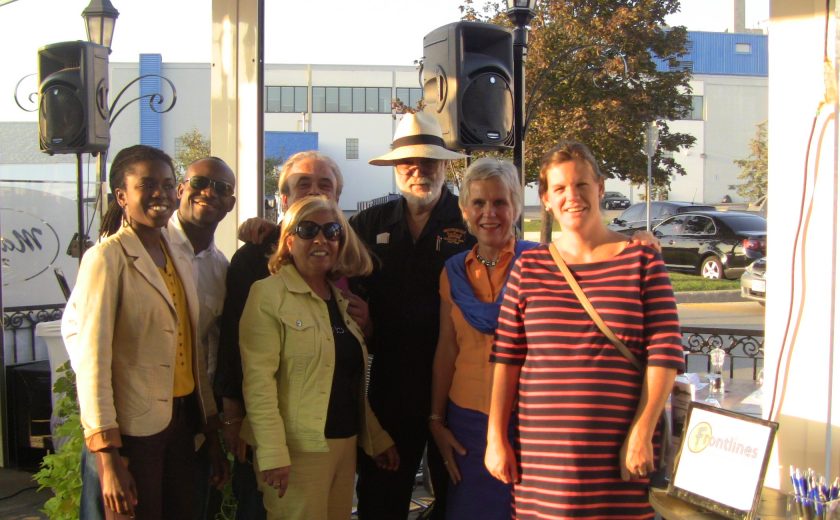 group of people smiling in front of two black speakers, blue sky, blue and white building in the background