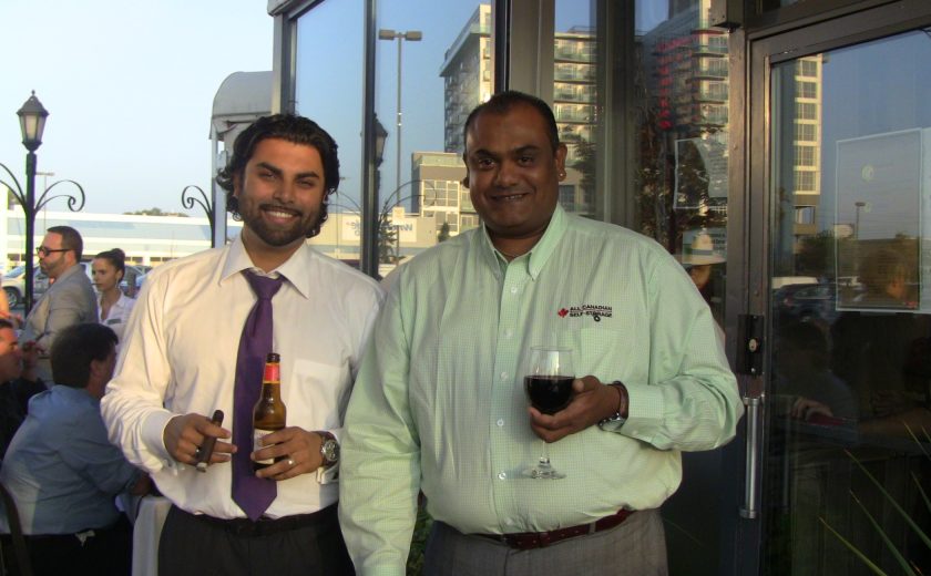 two men smiling, one wearing white shirt and purple tie, the other wearing a mint coloured shirt, standing in front of black exterior building, blue sky