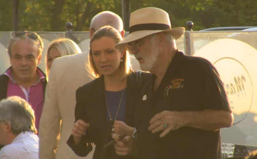 man wearing black and holding a cigar, woman talking wearing a blue dress and black blazer talking to the man, glass boarder surrounding the patio, green trees in the background