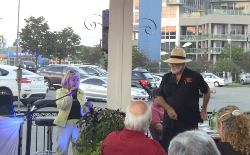 people sitting at tables listening to woman wearing a black shirt standing in front of a microphone, white pillar in the middle, buildings and parking lot in the background