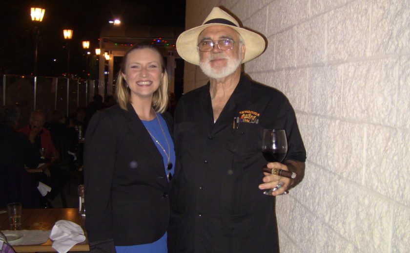 a women wearing a blue dress and black jacket standing next to a man wearing all black holding a cigar and wine glass, night sky, white brick wall on the side