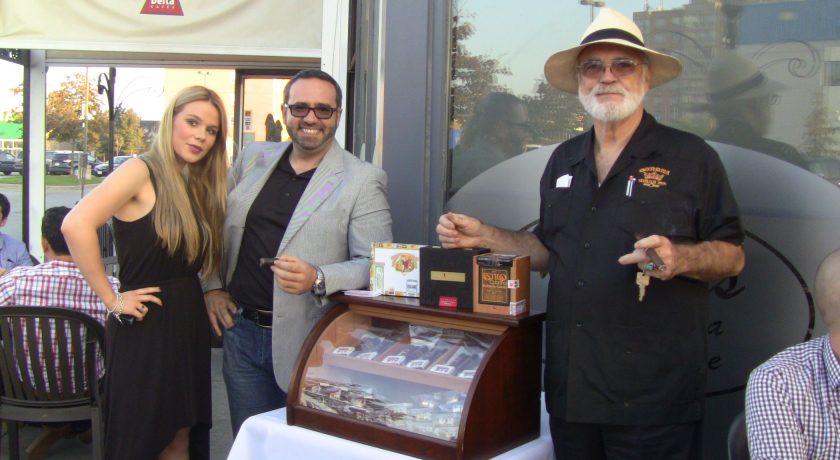 two men standing beside a mahogany case with a glass front displaying the cigars, woman standing beside the man on the left, case is on top of a white table, outside on a patio