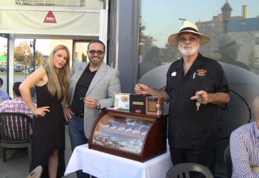 two men standing beside a mahogany case with a glass front displaying the cigars, woman standing beside the man on the left, case is on top of a white table, outside on a patio