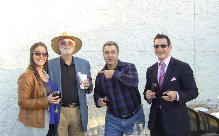 four people holding drinks standing behind a brown wooden table, white brick wall