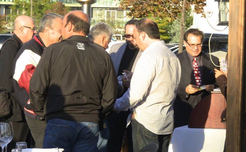 group of men gathering around each other outside, green trees and buildings in the background, in front of a restaurant with brown exterior