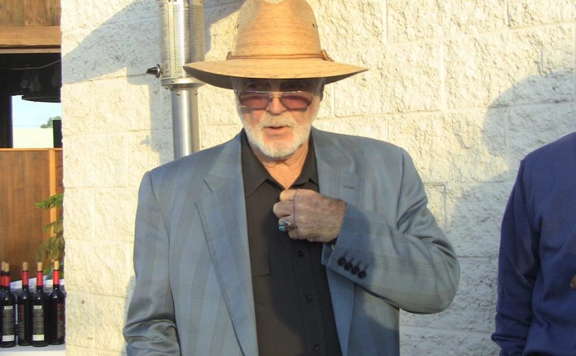 man wearing a light brown sun hat, black dress shirt, grey jacket and sunglasses against a white brick wall