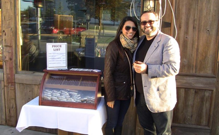 a man and a woman standing in front of a brown building with a large glass window, standing next to a mahogany case of cigars on a white square table