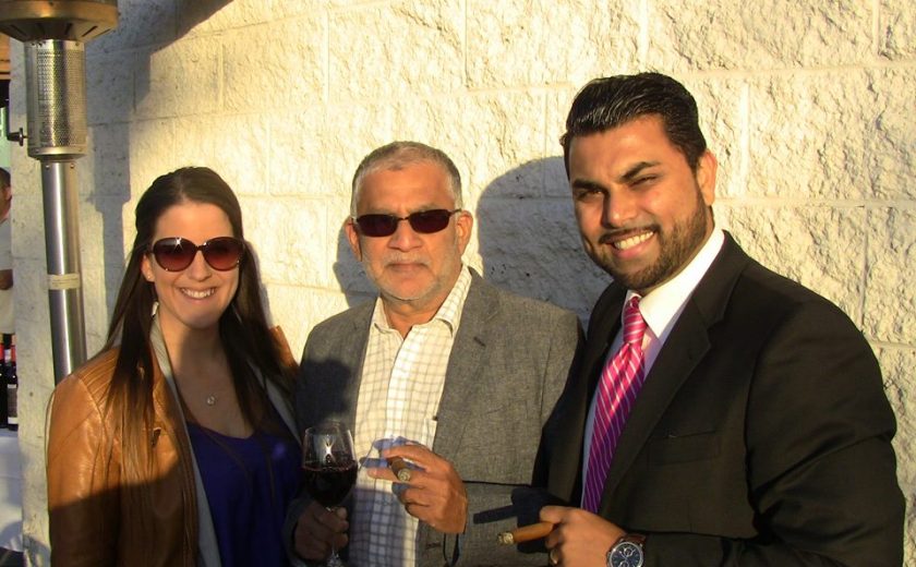 one woman wearing sunglasses and two men wearing dress shirts and jackets holding cigars, against a white brick wall, sunny day