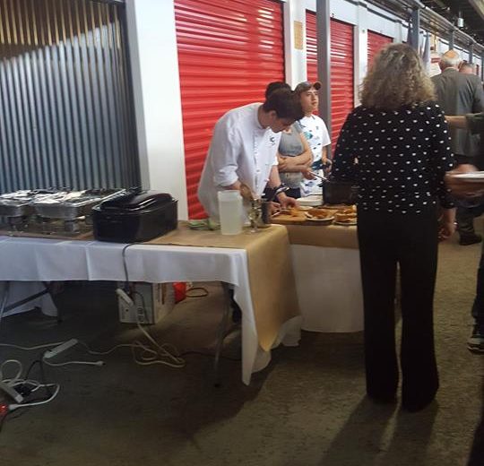 Flavours of Thorncliffe, people serving food in a storage facility, red storage units in the background