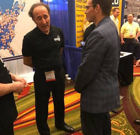 2017 Spring Conference & Trade Show, two men talking at a booth, Storage Auction Website backdrop in the background with a map