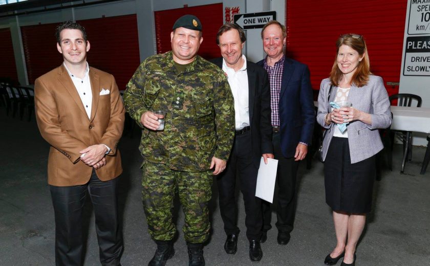 group of five nicely dressed up, one of the five is wearing a green uniform, red storage units in the background