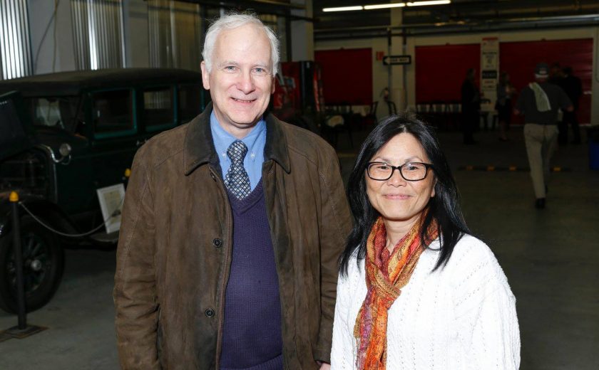 a man wearing a purple sweater vest, blue shirt and brown jacket standing next to woman wearing a white sweater with black glasses, old car green car on display, storage units with red doors in the background