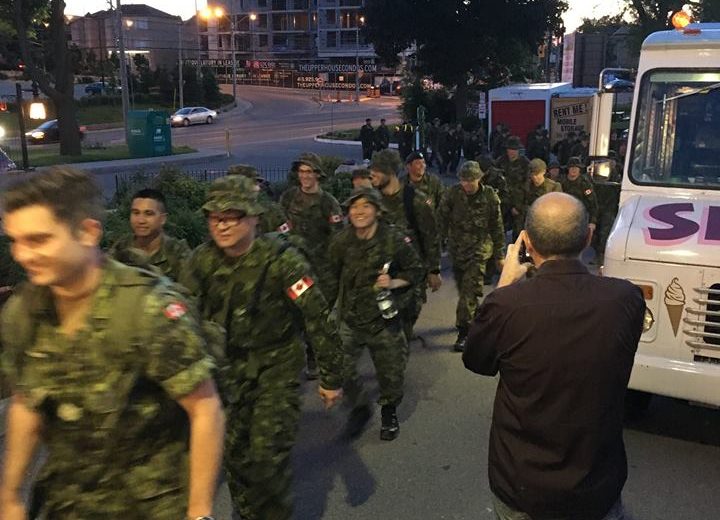 group of men wearing army uniforms walking outside in a straight line, white ice cream truck, building under construction in the background and dark green trees