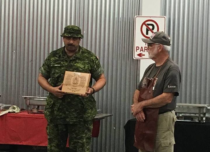 man holding wooden plaque wearing green army uniform, man wearing brown apron standing, silver wall with silver food containers on tables