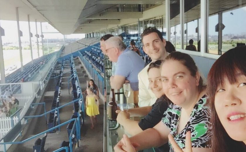 Woodbine Races, group selfie of 4 people outdoors at the race track, several rows of blue chairs in the background, under a cement roof