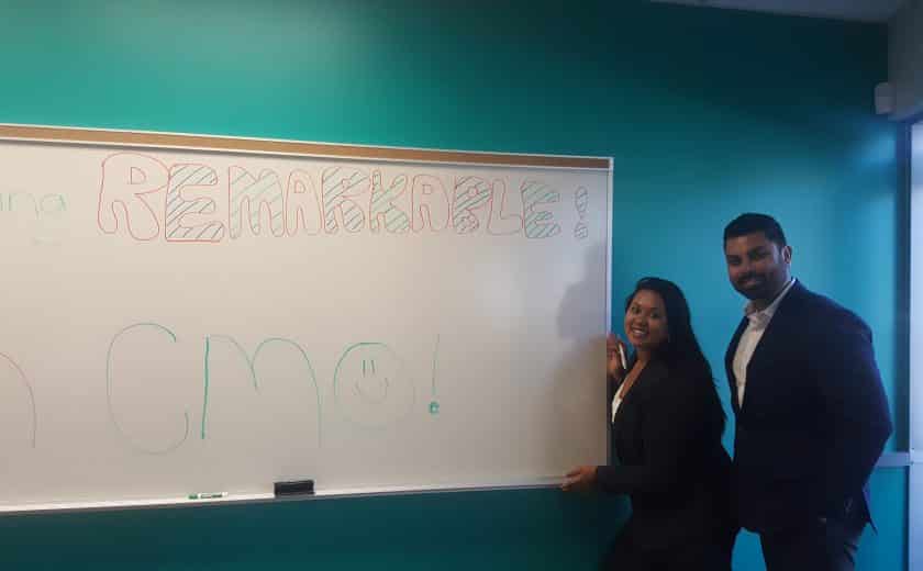 Man and woman wearing white dress shirt and black blazer standing on the right side of a large whiteboard in the Client Management Office, teal coloured wall as the background