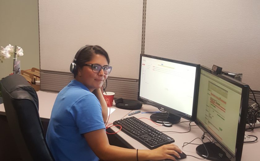 Woman wearing a blue t-shirt in front of two desktop computers in the client management office, yellow, red, white and purple balloon in the background