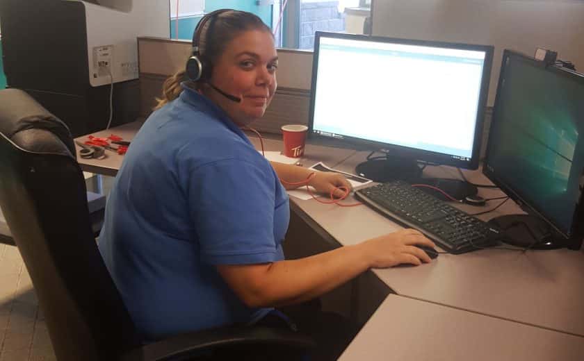 Woman wearing a blue t-shirt and headset in front of two computers, doors and windows in the background, white and green balloon in background