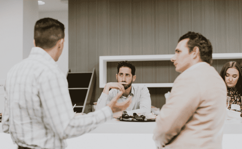 three men engaged in conversation in a white and brown room, woman sitting in the background