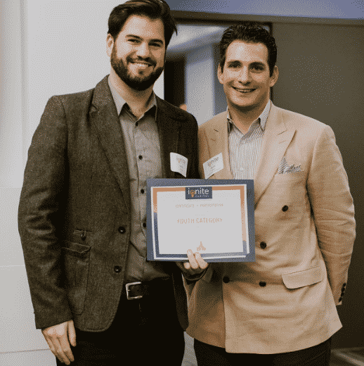 two men standing wearing a buttoned up shirt and a blazer with black pants smiling at camera holding ignite certificate