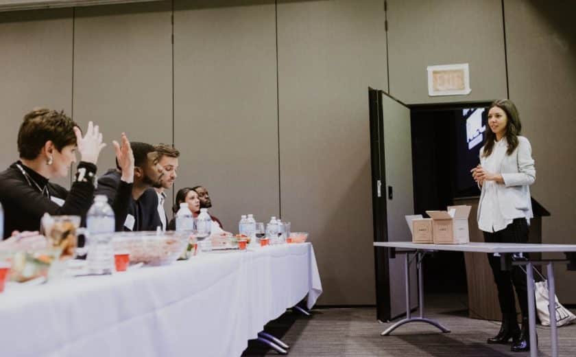 five people sitting at the judges panel behind a white long table listening to a young woman wearing white present at ignite capital pitch competition