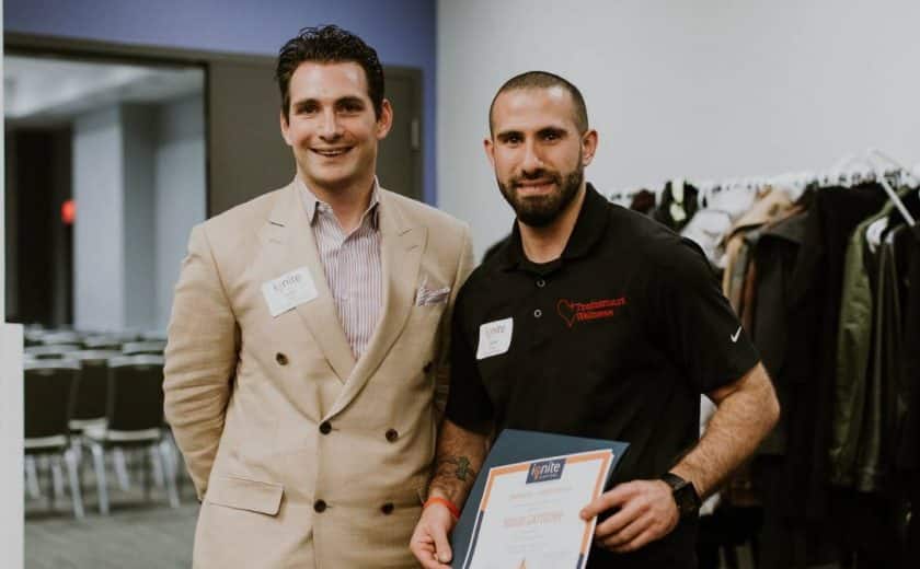 Two men standing smiling at the camera, one is wearing a striped shirt with light brown blazer jacket, the other man is wearing a black shirt holding an ignite capital certificate, jackets hanging on a rack in the background