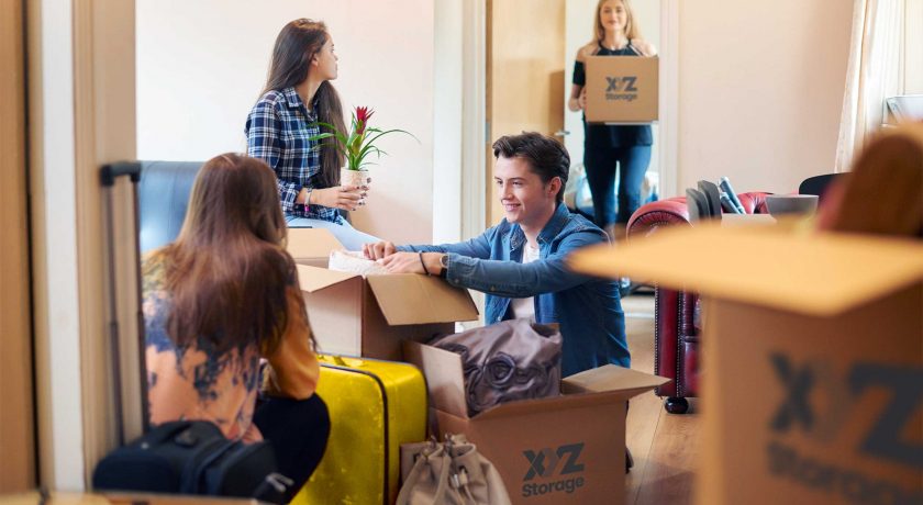 boy and girl on the floor packing their apartment with XYZ Storage moving boxes, one girl sitting on a couch holding a plant, another girl standing holding an XYZ Storage moving box