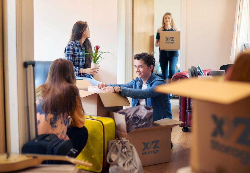 boy and girl on the floor packing their apartment with XYZ Storage moving boxes, one girl sitting on a couch holding a plant, another girl standing holding an XYZ Storage moving box