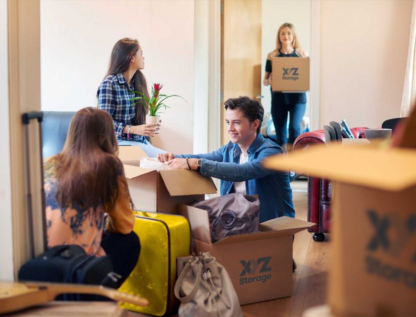 boy and girl on the floor packing their apartment with XYZ Storage moving boxes, one girl sitting on a couch holding a plant, another girl standing holding an XYZ Storage moving box