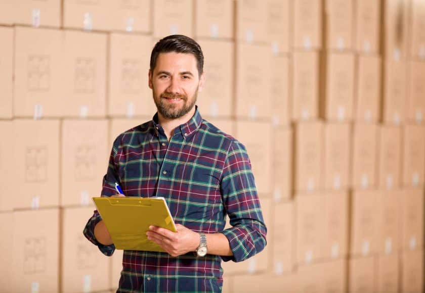 man wearing a colourful plaid shirt holding a yellow clipboard a blue and white pen, wall of brown boxes in the background