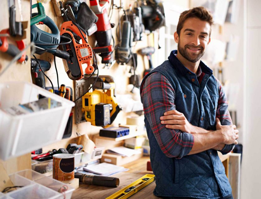 man wearing a blue and red plaid shirt and a blue vest smiling, leaning against a desk with a bunch of tools hanging on the wall