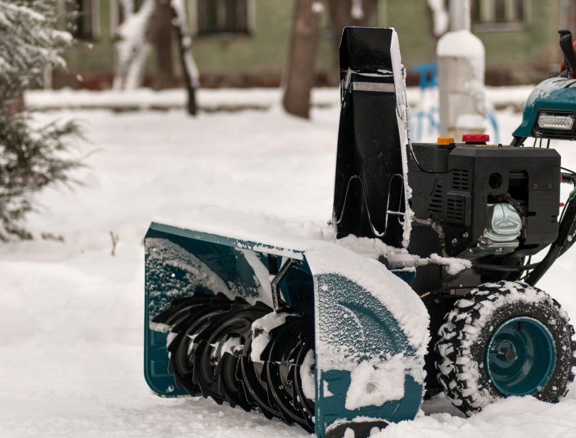 teal and black snow plower plowing white snow, green building in the background, tree on the side