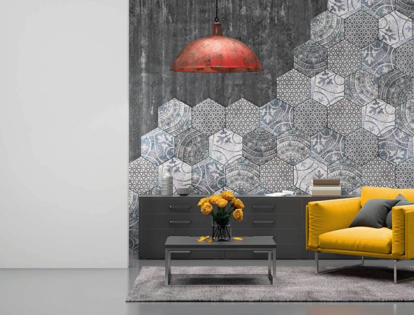 grey room with a hexagon designed wall, long dark grey dresser against the wall with brown and white book on top, yellow armchair with grey pillows, coffee table with yellow flowers, a red and brown ceiling light