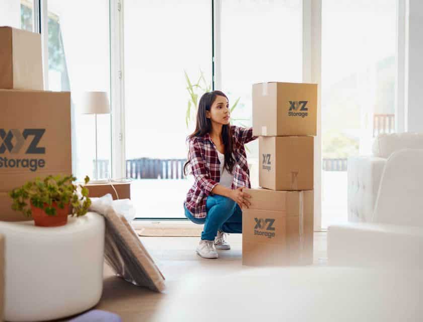 young woman wearing a plaid shirt with blue jeans and white shoes, packing her belongings using XYZ Storage boxes