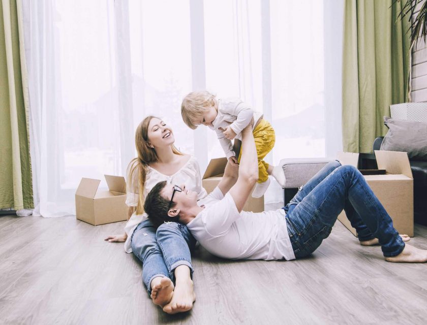 family of three laying on the floor in a house surrounded by brown boxes and a big window in the background