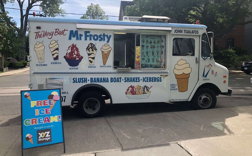 white ice cream truck with colourful pictures of ice cream beside a grey sidewalk, green trees in the background, colourful sign in front saying free ice cream brought to you by XYZ Storage