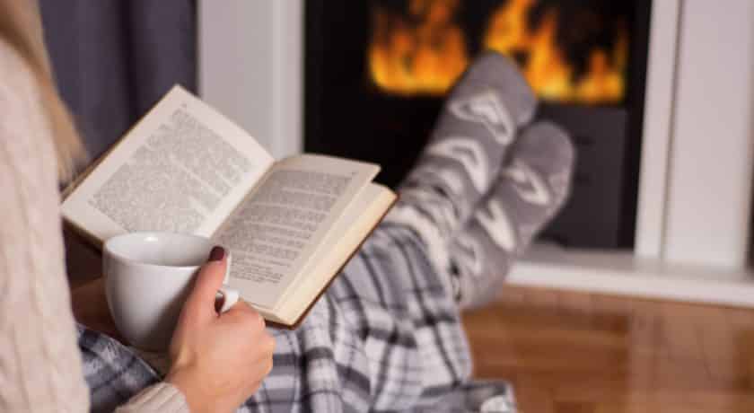 Beautiful young woman in front of the fireplace reading book and warming feet on fire and legs are covered with blanket, in hand holds cup of hot tea. Winter and cold weather concept at home. Close up, selective focus