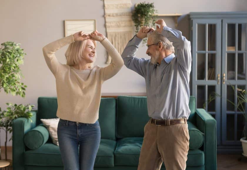 Funny sixty years old couple in love having fun at home, elderly spouses standing raised hands dancing feels carefree healthy enjoy active weekend, do exercises well being of retirees people concept