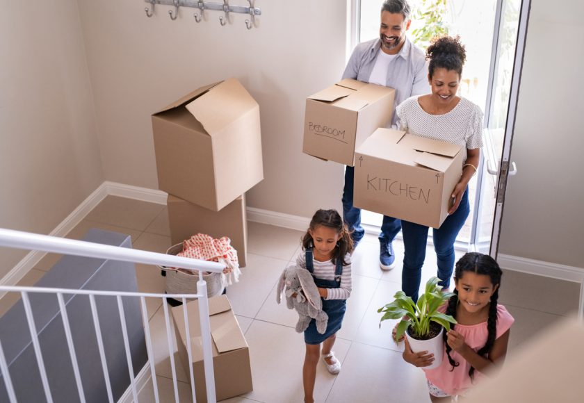 Ethnic family with two children carrying boxes and plant in new home on moving day. High angle view of happy smiling daughters helping mother and father with cardboard boxes in new house. Top view of excited kids having fun walking up stairs running to their rooms while parents holding boxes.